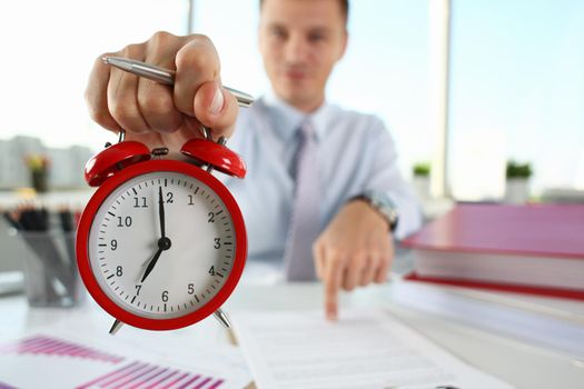 Man hand on red alarm clock stands at desk in office showing seven o'clock in morning or in evening AM PM. Overtime work deadline concept