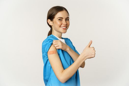 Smiling nurse, doctor in scrubs, medical worker showing her vaccinated shoulder with patch, thumbs up, recommending vaccinate from coronavirus, white background.