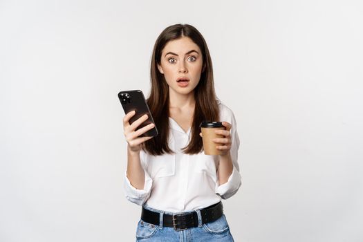 Portrait of young woman looking surprised after checking mobile phone, holding takeout cup of coffee, standing over white background.