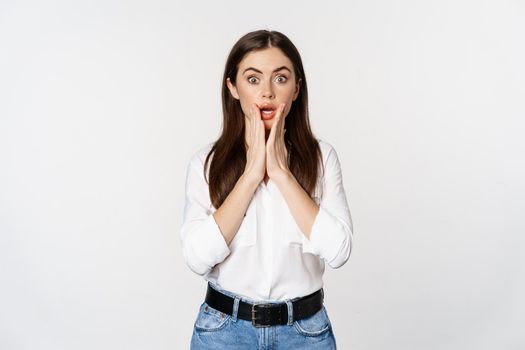 Surprised brunette woman looking amazed, seeing smth awesome, standing in blouse and jeans over white background.