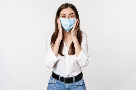 Surprised woman in face medical mask looking amazed, checking out awesome news, big reveal, standing over white background.