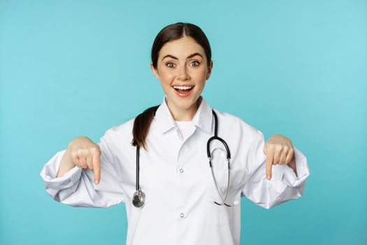 Enthusiastic healthcare worker, young woman doctor in white coat, pointing fingers down and smiling, showing pharmacy advertisement, medical promo, blue background.