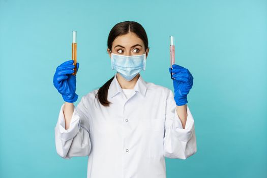 Image of young woman doctor, lab worker doing research, holding test tubes, wearing medical face mask and rubber gloves, blue background.