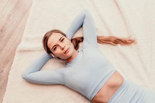 Top view portrait of relaxed woman listening to music with headphones lying on carpet at home. She wears a blue suit with long hair pulled back into a ponytail