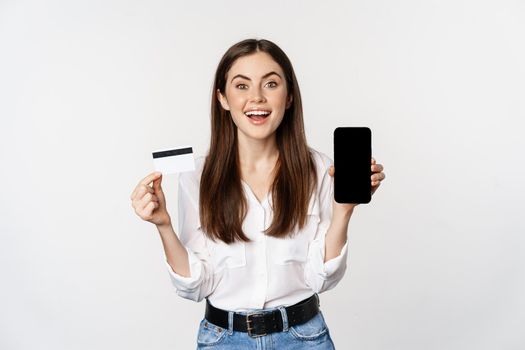 Happy woman showing credit card and smartphone screen, concept of online shopping, buying in app, standing over white background.