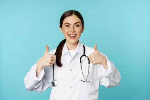 Happy woman doctor, medical worker in white coat, showing thumbs up in approval, like something, praise, standing over blue background.