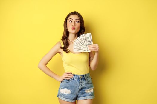Shopping, credit and money concept. Young brunette woman showing cash and smiling pleased, standing over yellow background.