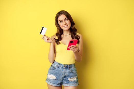 Online shopping. Stylish brunette woman holding smartphone and credit card, paying in app, using mobile phone application, buying smth, standing over yellow background.