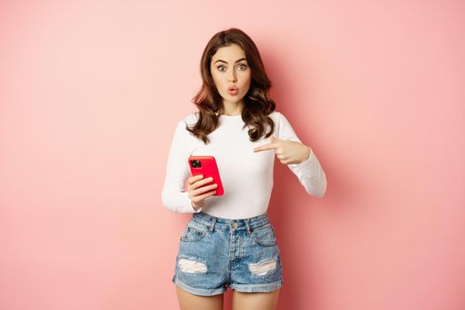 Cute girl pointing finger at smartphone with curious face expression, have you seen this gesture, showing online shopping offer on mobile phone app, pink background.