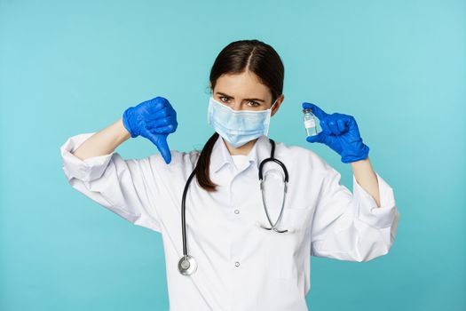 Sad and disappointed young doctor, woman showing vaccine and dislike, thumbs down gesture, disapprove medical drug, standing over blue background.