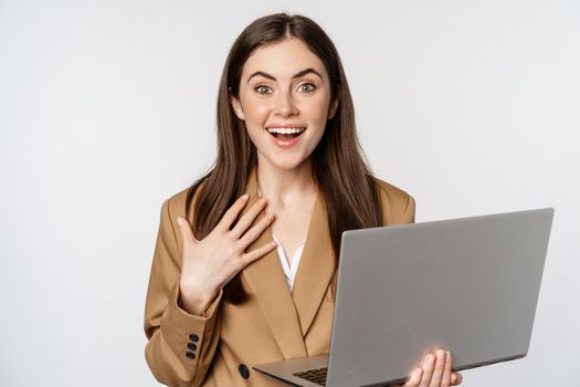 Surprised and excited businesswoman holding laptop, reacting amazed at smth awesome, standing in suit over white background.