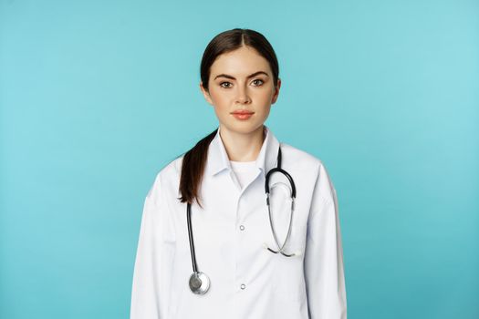 Medical staff and doctors concept. Young smiling female doctor, healthcare worker in white coat and stethoscope, looking confident, waiting patients, blue background.