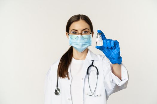 Portrait of young woman, doctor in medical face mask and uniform, showing vaccine, covid-19 vaccination campaign, standing over white background.