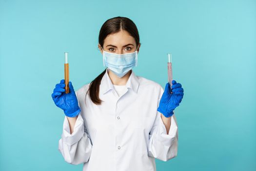 Image of young woman doctor, lab worker doing research, holding test tubes, wearing medical face mask and rubber gloves, blue background.