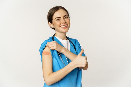 Happy smiling medical worker, doctor or nurse showing her shoulder after vaccination, covid-19 vaccine and thumbs up satisfied, standing over white background.