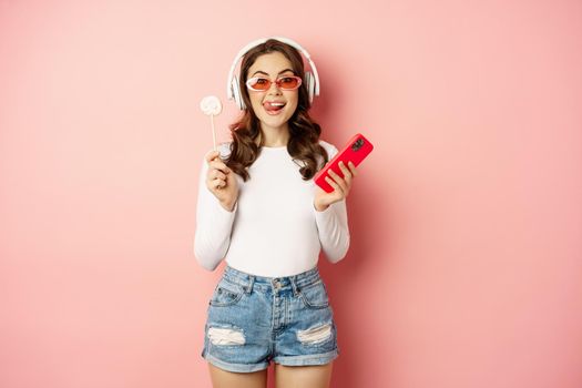Stylish brunette woman dancing in headphones with mobile phone, listening music in earphones, licking lolipop, standing over pink background.