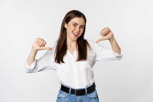 Portrait of young woman pointing at herself, self-promoting, standing over white background. Copy space