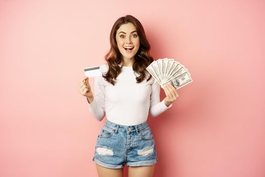 Credit, money and loans concept. Happy beautiful girl holding credit card and cash, looking satisfied, thinking of shopping, standing over pink background.