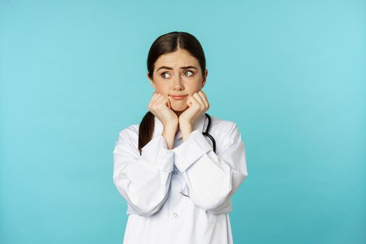 Scared and worried woman doctor in white coat, looking anxious and insecure, shaking from fear, standing over torquoise background.