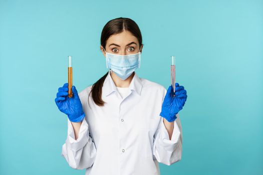 Image of young woman doctor, lab worker doing research, holding test tubes, wearing medical face mask and rubber gloves, blue background.