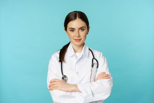 Medical staff and doctors concept. Young smiling female doctor, healthcare worker in white coat and stethoscope, looking confident, waiting patients, blue background.