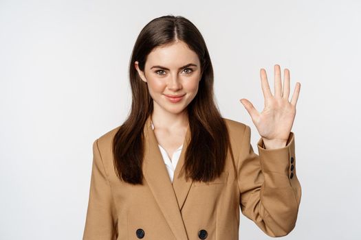 Portrait of corporate woman, saleswoman showing number five fingers and smiling, standing in suit over white background.