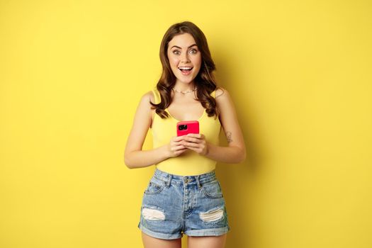 Cellular technology and online shopping concept. Enthusiastic pretty girl standing with smartphone and smiling, standing against yellow background.