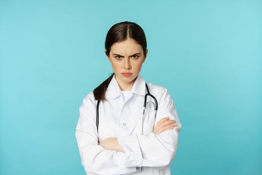 Angry doctor, female medical worker looking offended, cross arms on chest, frowning and staring annoyed at camera, standing over blue background.