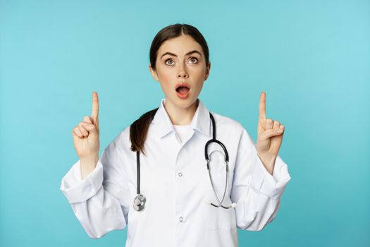 Enthusiastic medical worker, young woman doctor in white coat, stethoscope, showing advertisement, pointing fingers up, standing over torquoise background.