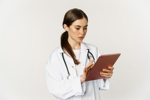 Woman doctor looking concerned at digital tablet, reading with worried face expression, wearing white coat, standing over white background.