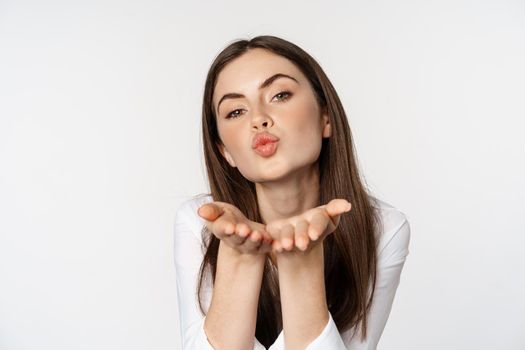 Feminine woman sending air kiss at camera, coquettish flirty pose, kissing, standing over white background. Copy space