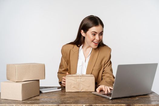 Smiling happy businesswoman working in her office with packages delivery, process orders on her laptop website, sending goods to clients, white background.