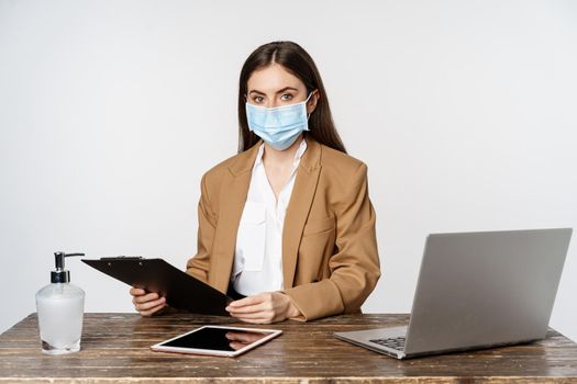 Covid, pandemic and office workplace concept. Portrait of corporate woman in medical face mask from coronavirus, working, standing over white background.