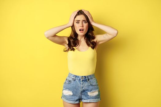 Troubled and shocked woman holding hands on head, panicking, feeling distressed and anxious, having problem, standing over yellow background.