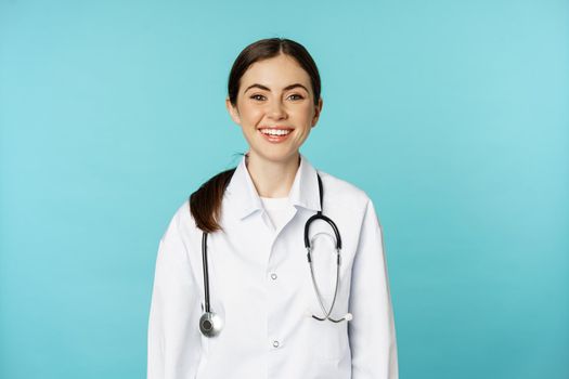 Smiling healthy medical worker, young woman doctor looking happy, standing in white coat and stethoscope against blue background.