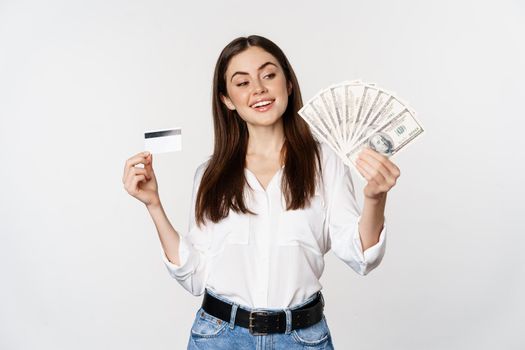 Happy modern woman holding money and credit card in hands, smiling pleased, standing over white background. Copy space