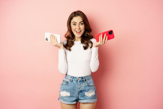 Technology, spring promo concept. Stylish glamour woman holding two smartphones, mobile phones in both hands, smiling pleased, buying new cellphone, pink background.