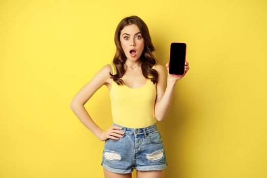 Stylish smiling woman showing mobile phone screen, app interface on smartphone, standing in tank top and shorts against yellow background.
