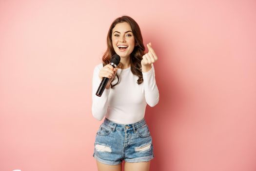 Party girl. Happy young woman singing in microphone, performing song, having fun at event, standing over pink background.