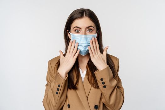 Workaplce and pandemic concept. Shocked business woman in face medical mask, gasping, looking startled and concerned at camera, standing over white background.