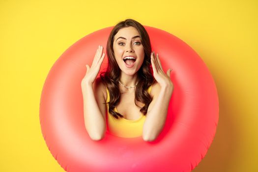 Beautiful coquettish woman inside pink swimming ring, posing on summer vacation, beach holiday concept, standing against yellow background.
