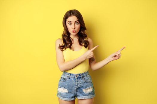 Beautiful young woman pointing fingers at banner, logo or advertisement, smiling amazed, standing in summer clothes, yellow background.