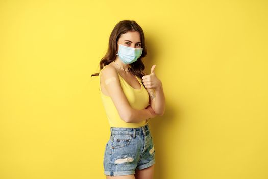 Portrait of young woman got vaccinated from covid19, flu, wearing face mask, showing thumbs up, girl has patch on shoulder from vaccination, standing over yellow background.