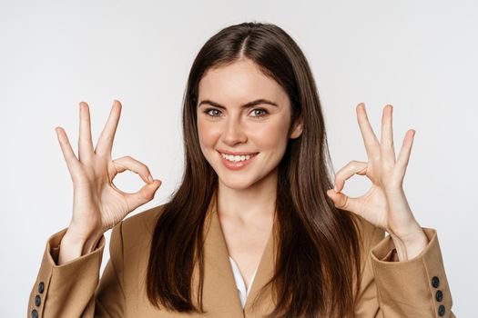 Close up portrait of confident businesswoman smiling, showing okay signs, recommending company, approve and like smth good, standing over white background.