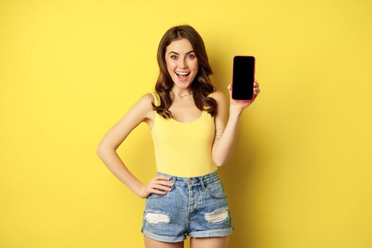 Stylish smiling woman showing mobile phone screen, app interface on smartphone, standing in tank top and shorts against yellow background.