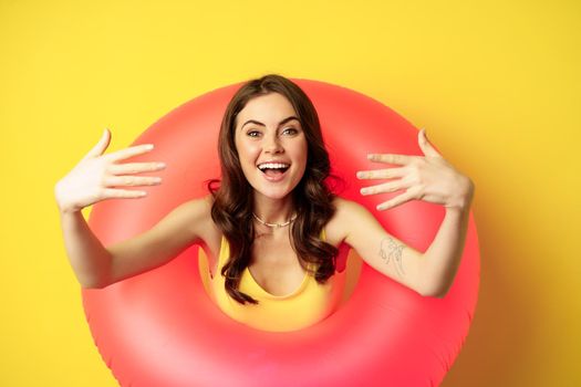 Close up portrait of beautiful happy woman inside pink swimming ring, luring, inviting people on summer vacation, beach party, standing over yellow background.