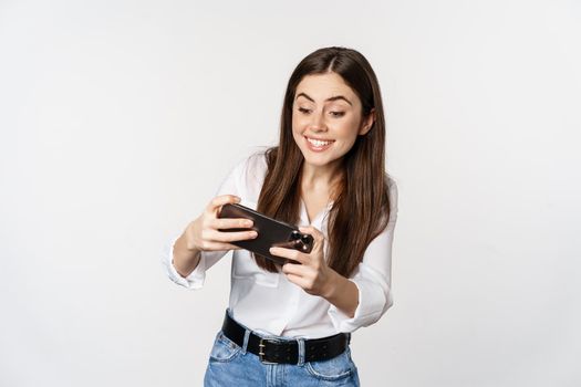 Happy brunette woman playing mobile video game, smiling and looking at screen excited, standing over white background.