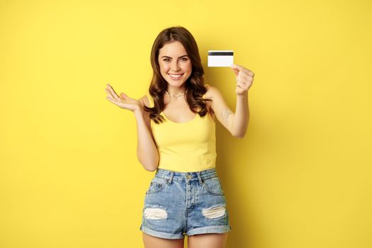 Portrait of brunette girl showing her credit card and smiling, wearing summer clothes, concept of bank, money and shopping, standing over yellow background.