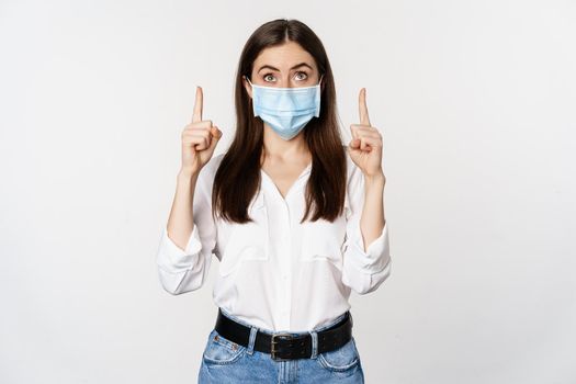 Portrait of corporate woman in face medical mask from coronavirus, pointing fingers up, showing advertisement, white background.