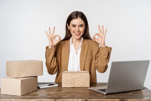 Woman small business owner, sitting at table with laptop, packing boxes, processing clients orders, showing okay, ok sign and smiling, posing against white background.